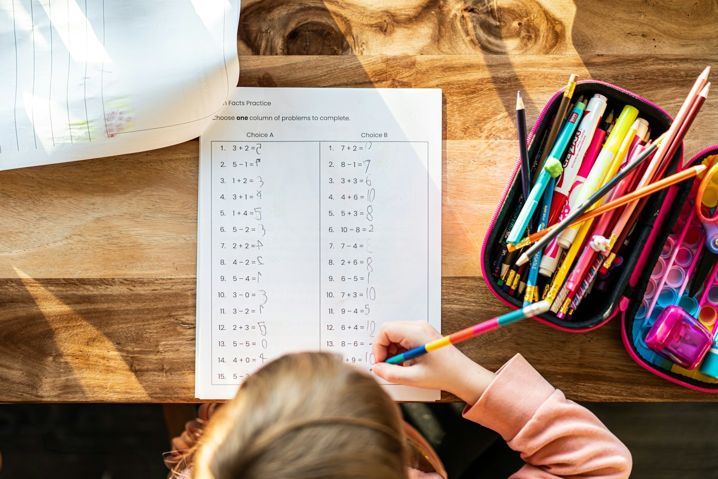 Dieťa vypĺňa matematický pracovný list pri drevenom stole.